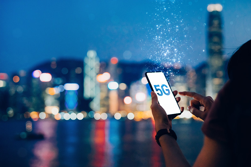 晚上，一名年輕女子在維多利亞港的海濱長廊上使用智能手機，背景是香港標志性的城市天際線，5G通信的概念被分解成輕粒子圖片素材