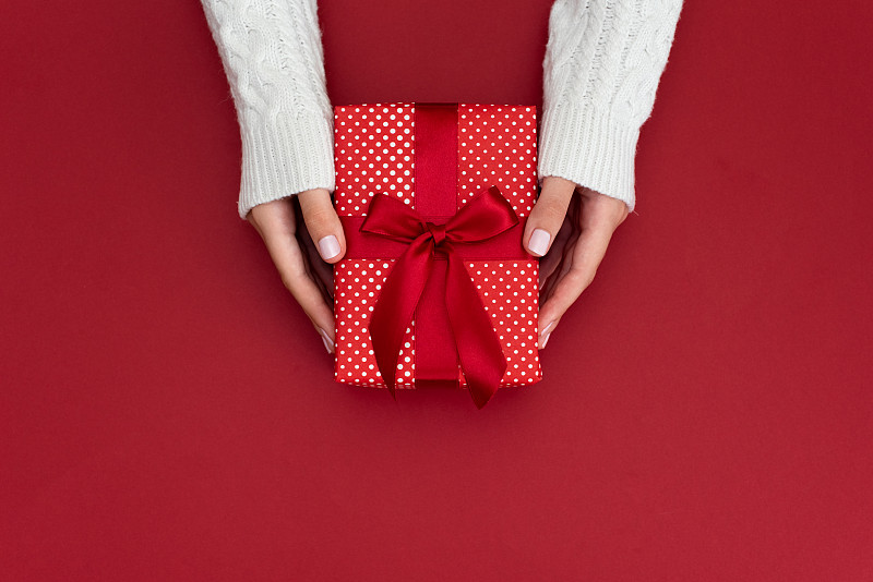 女人的手拿著禮物盒與紅色蝴蝶結在柔和的粉紅色背景與五彩紙屑。平的風格。圖片素材