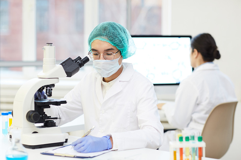 嚴肅年輕的亞洲實驗室研究員，穿著無菌工作服，坐在書桌前，用顯微鏡記錄樣品圖片素材