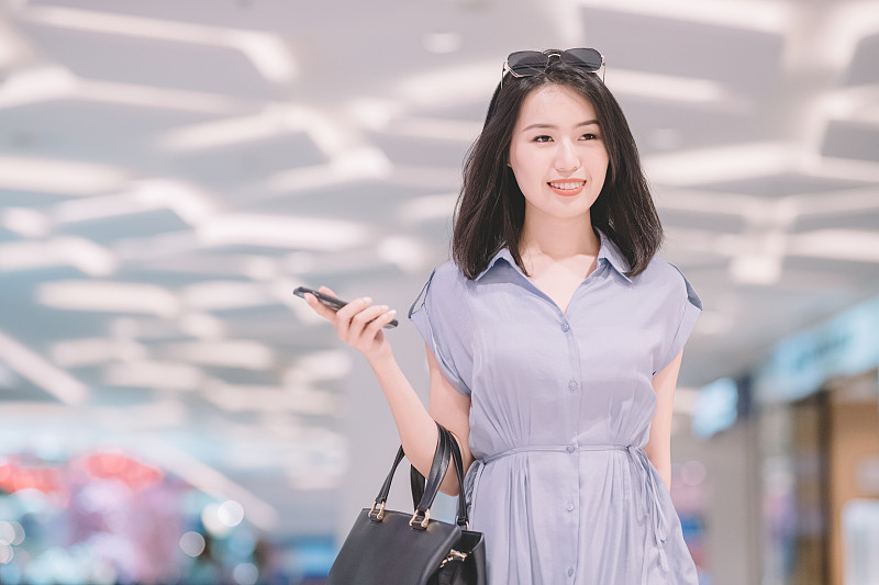 an asian chinese female walking in the shopping mall shopping holding smart phone and purse圖片素材