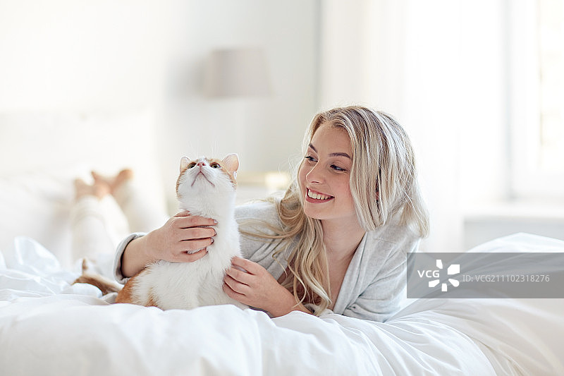 宠物，早上，舒适，休息和人的概念-快乐的年轻女人与猫在床上在家图片素材