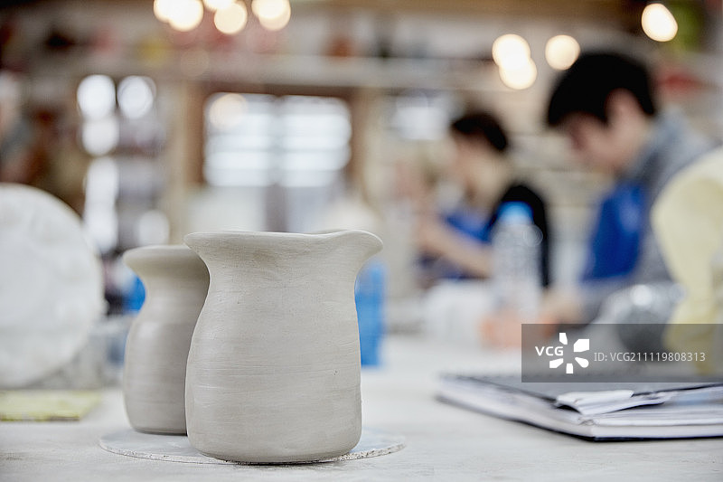 前景是两个陶罐。一节陶艺课正在进行，人们坐在陶艺工作室的工作台上。图片素材