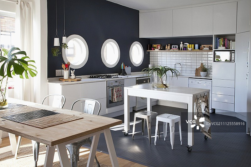 在白色的厨房里，黑色马赛克地板上的小脚轮桌，黑色的强调墙，舷窗和前景支架上的木桌图片素材