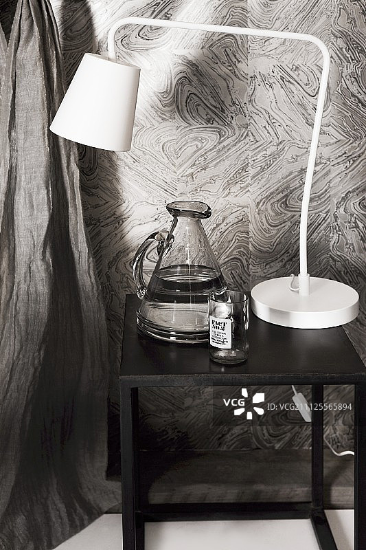 黑色的边桌上放着一盏复古的白色台灯和一壶水，映衬着灰色图案的墙纸图片素材