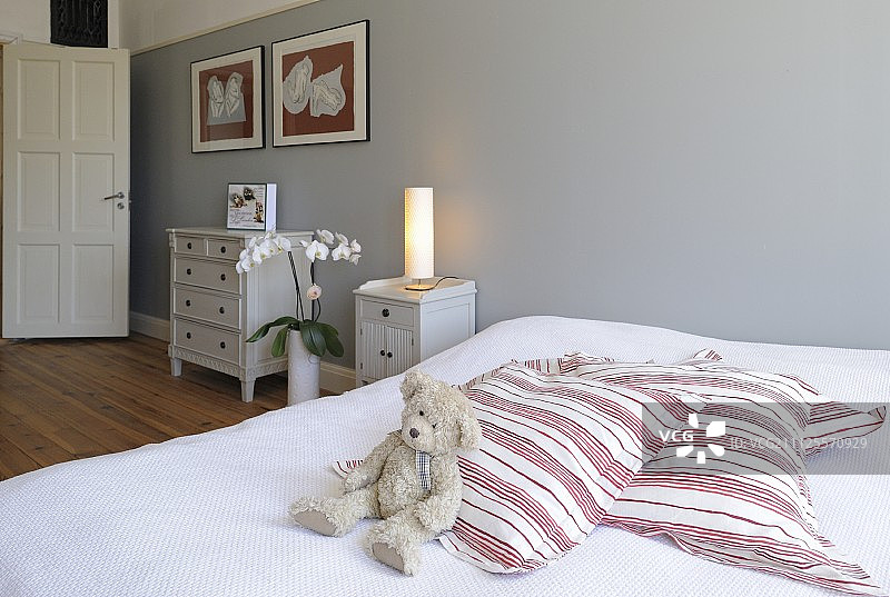 双人床上灰墙边放着泰迪熊和条纹软垫;背景是乡村别墅风格的抽屉柜图片素材