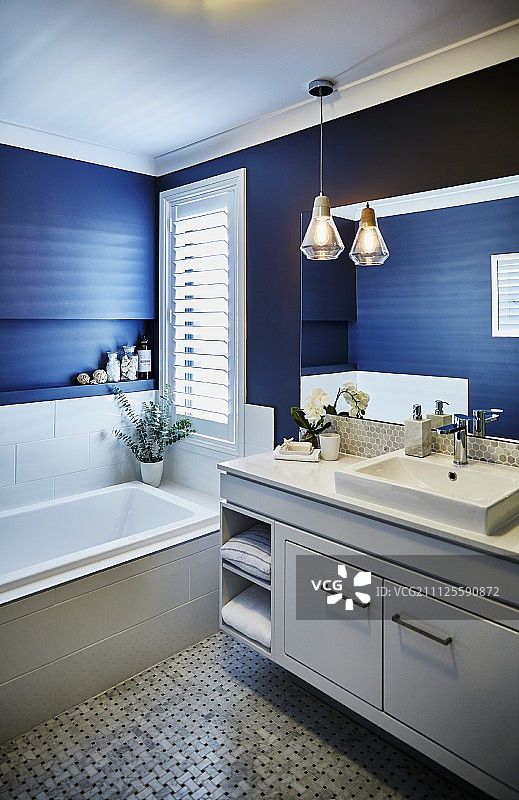在蓝色的浴室里，有一个白色的盥洗柜，旁边是安装好的浴缸，旁边是窗户图片素材