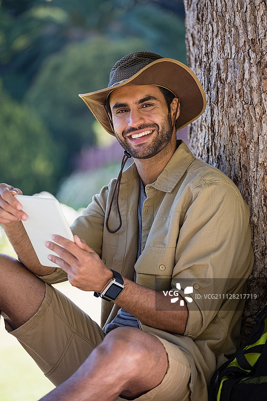 在森林里的一棵树旁休息时，微笑的男人在使用数字平板电脑图片素材