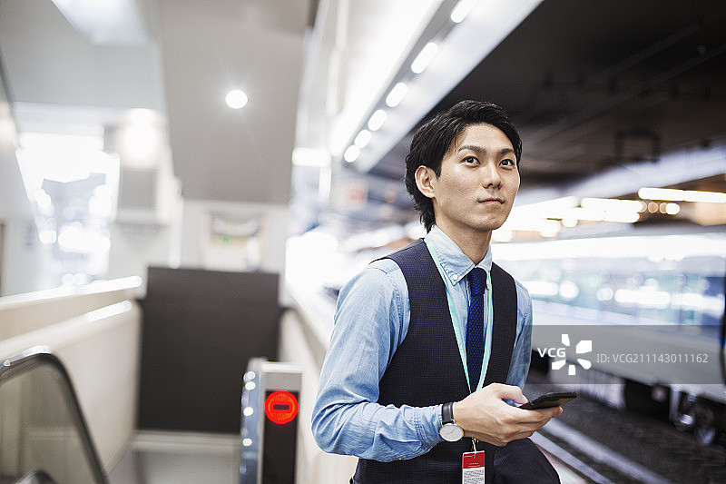 商人穿着蓝色衬衫和背心站在火车站的月台上，拿着手机。图片素材