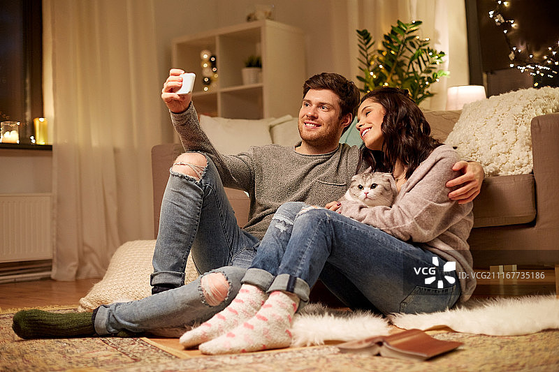 休闲，科技和人的概念-幸福的夫妇与猫在家里用智能手机自拍。幸福的夫妇在家里用智能手机自拍图片素材