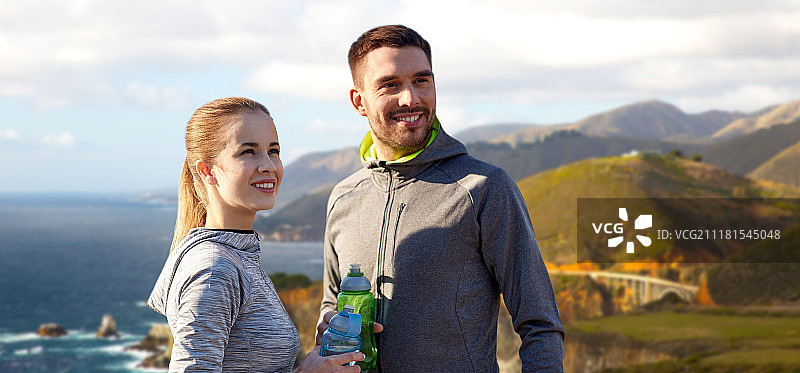 健身，运动和人的概念-微笑的夫妇与瓶装水在比克斯比溪桥在大的海岸加利福尼亚背景。两个运动员带着水越过大海岸。两个运动员带着水越过大海岸图片素材