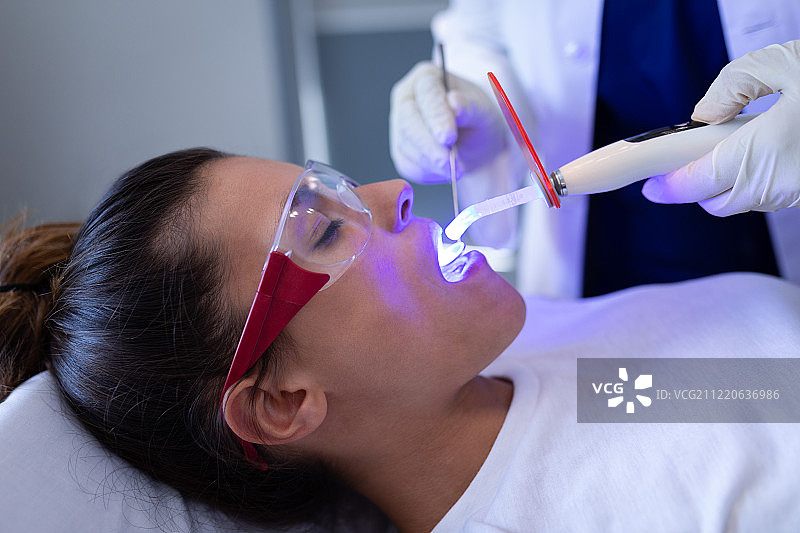 牙科医生在诊所用牙科治疗灯检查白种人病人的侧视图，病人戴着安全眼镜并张开她的嘴。图片素材