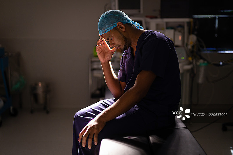 一个紧张的非裔美国男性外科医生坐在他的头在他的手在医院的手术室在光线下图片素材