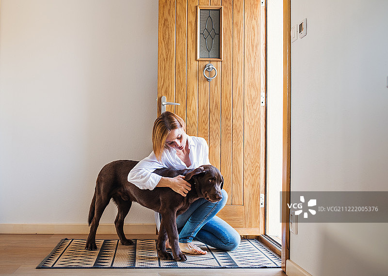 一个年轻的女人坐在家里的门旁的地板上，玩着一只狗。图片素材
