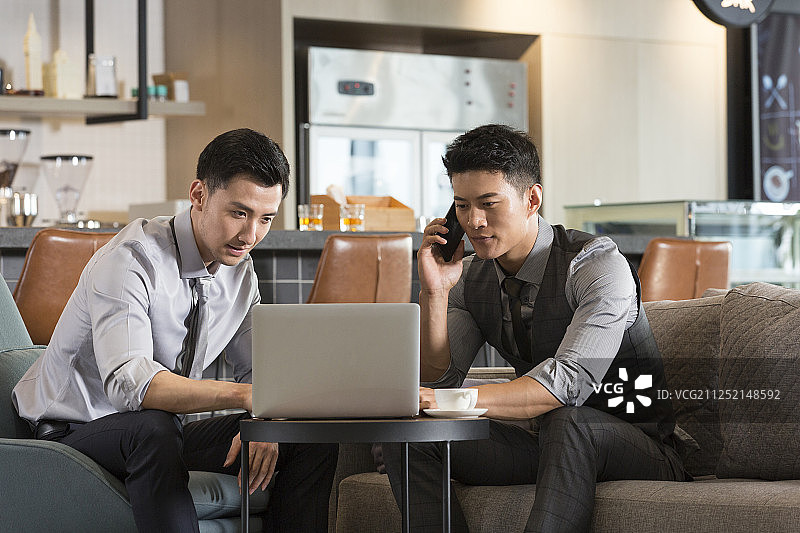 两个成功男商务人士在酒店咖啡馆使用笔记本电脑打电话谈论工作图片素材