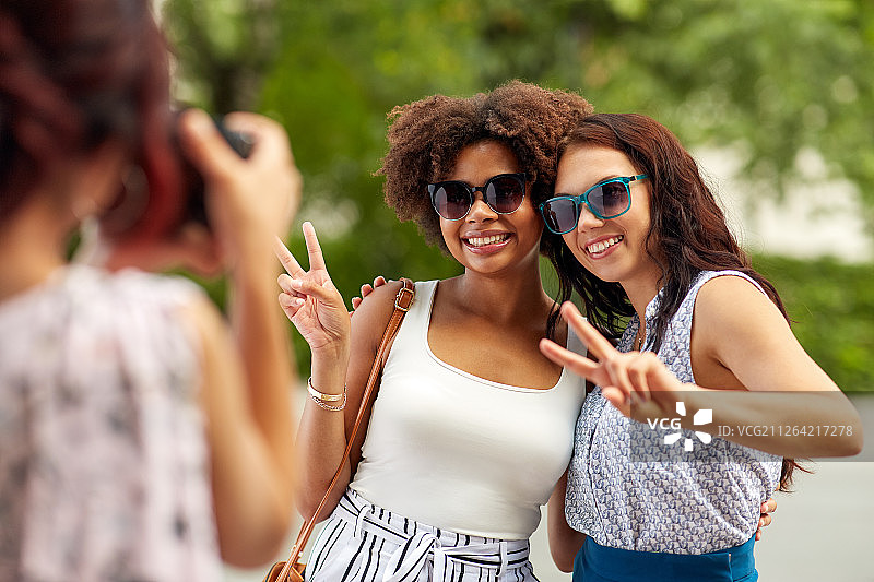 女性的友谊，科技和人-女人用相机拍摄她的朋友在夏季公园展示和平的手势。一个女人在夏天的公园里给她的朋友拍照图片素材