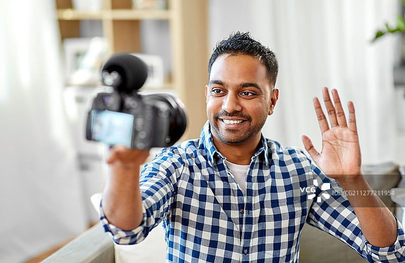 博客，视频博客和人的概念-微笑的印度男性视频博客与相机视频博客和挥手在家。男性视频博主用相机在家写博客图片素材