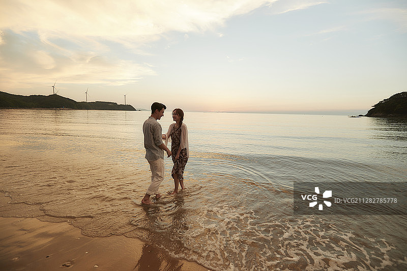 在日落的海滩上，一对夫妇手牵着手在水里泡脚图片素材