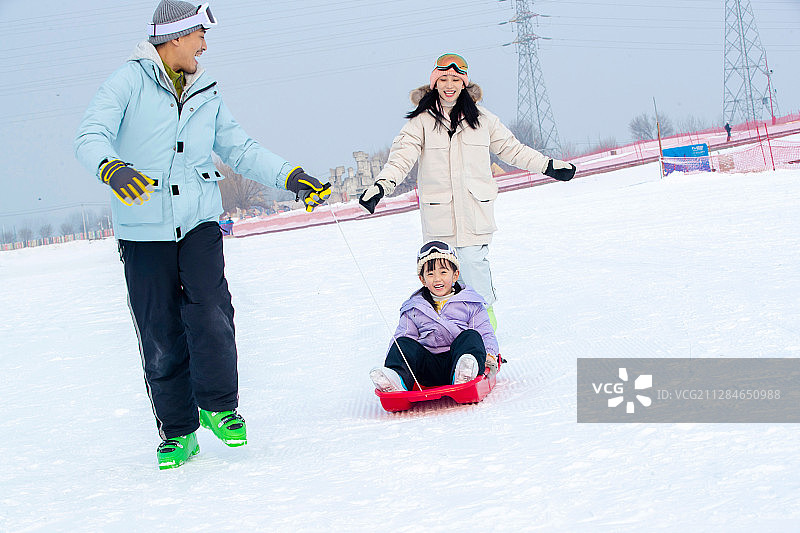 滑雪场上父母和坐在雪上滑板的女儿玩耍图片素材