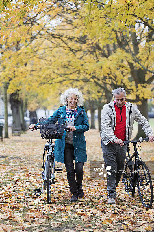 一对老年夫妇在秋天的公园里的树木和树叶之间散步自行车图片素材
