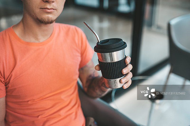泰国清迈，一名身穿橙色t恤的白人男子在咖啡馆里拿着可重复使用的咖啡杯图片素材