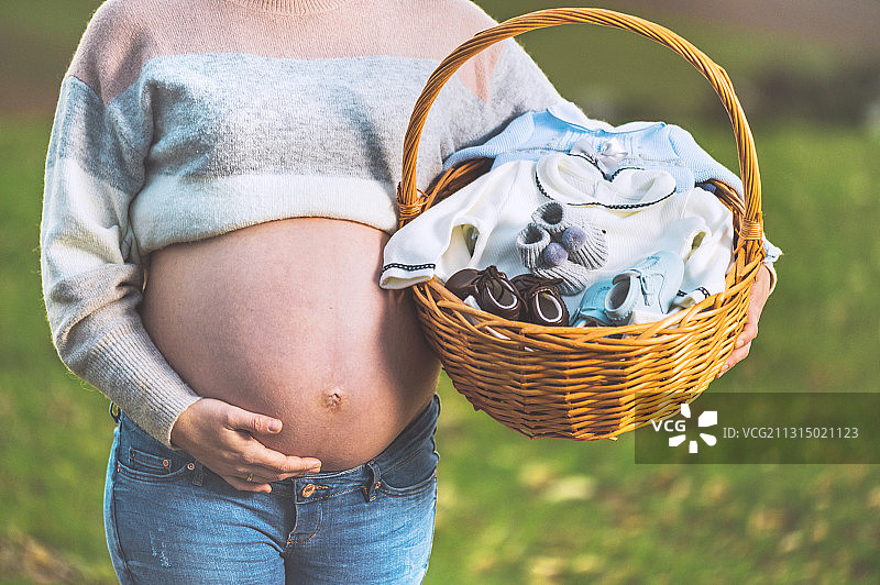 孕妇腹部抱着婴儿衣服篮子站在公园里图片素材