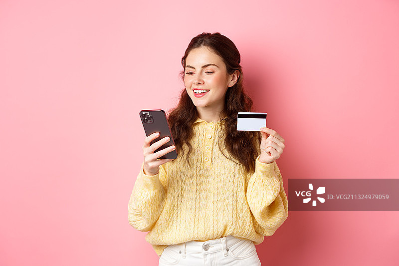 微笑的年轻女子用手机，同时拿着信用卡在粉红色的背景下图片素材