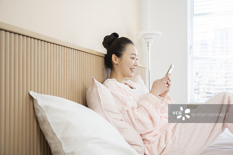 微笑快乐的亚洲女人穿着睡衣坐在床上用智能手机图片素材