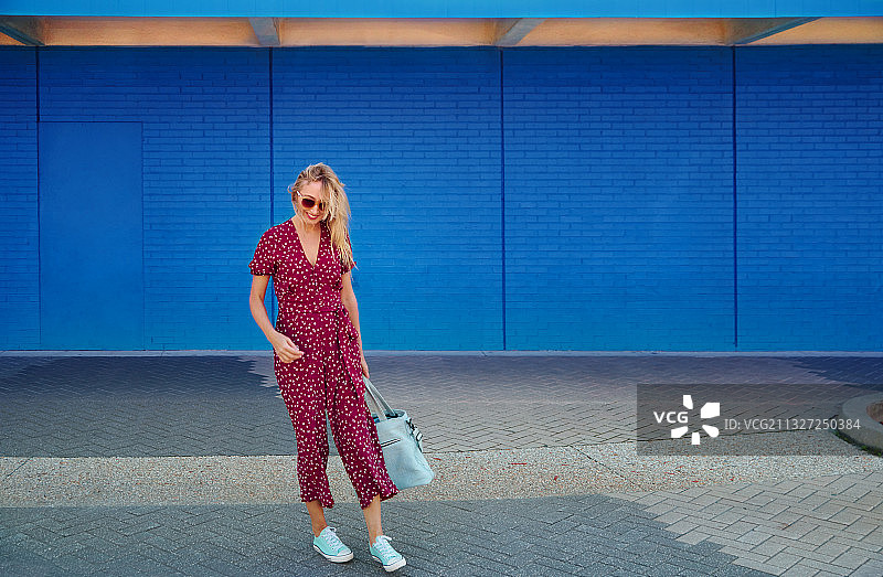 金发女人穿着紫红色的连身衣，戴着太阳镜，背着蓝色的墙走着，拎着蓝色的大手提袋图片素材