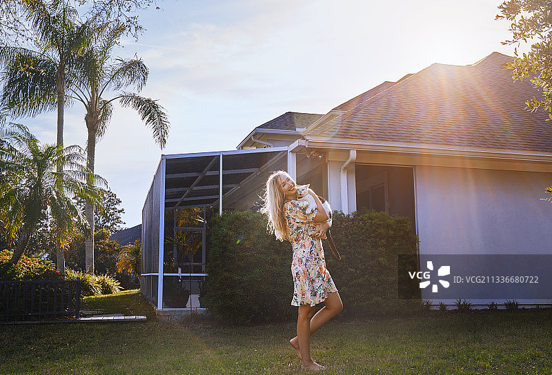 日落时分，微笑的金发女子与她的猫朋友在她的房子旁边拥抱图片素材