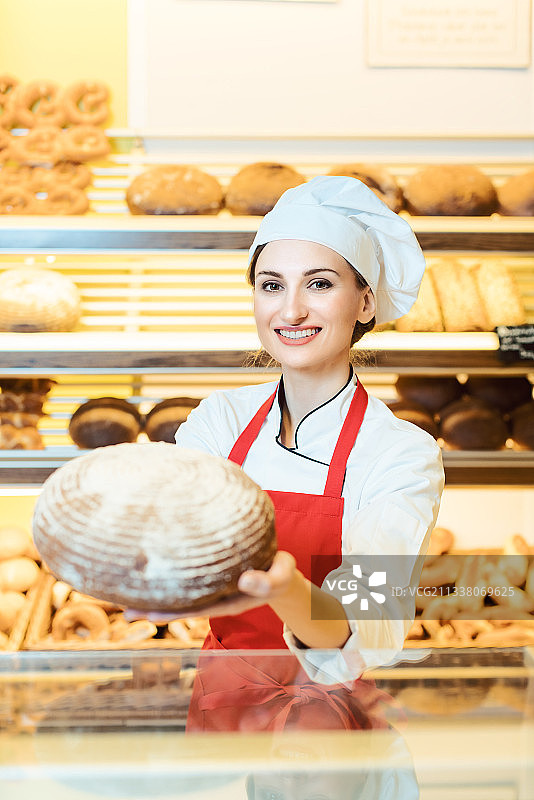 在面包店里，戴着围裙的女售货员正对着镜头展示新鲜的面包图片素材
