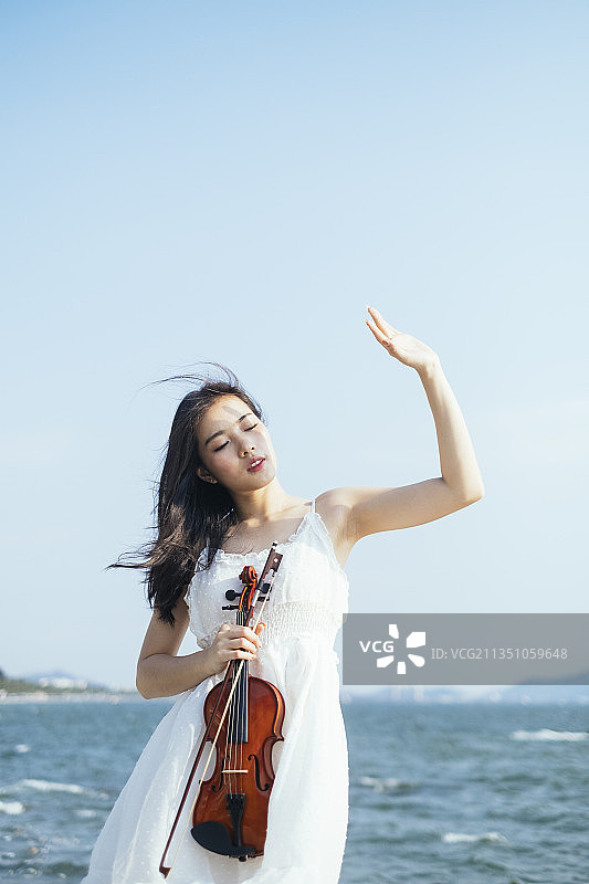 有风的日子，身穿白裙的长发女子站在海边，举起手来挡住阳光。图片素材