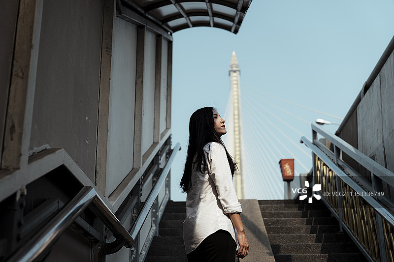 低角的黑头长发身穿白衬衫的女子站在阳光下的桥楼梯上，望着天空。图片素材