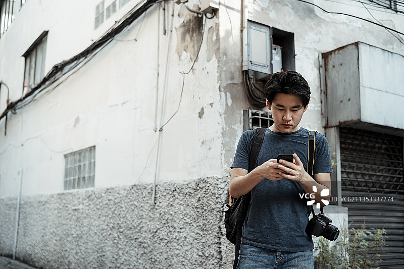 年轻英俊的亚洲摄影师男子使用智能手机在曼谷的街道小巷。图片素材