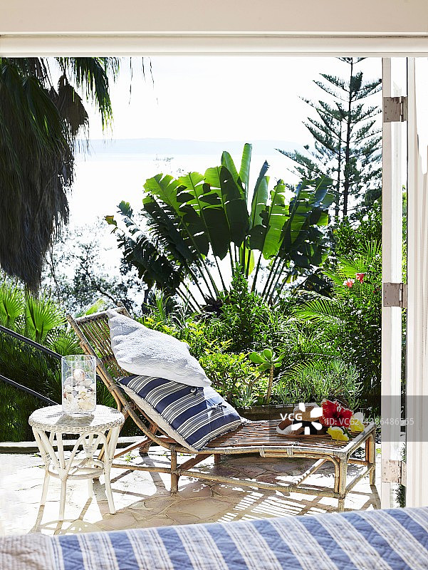竹制阳光躺椅和白色柳条桌在阳光明媚的露台上，海景和热带植物图片素材
