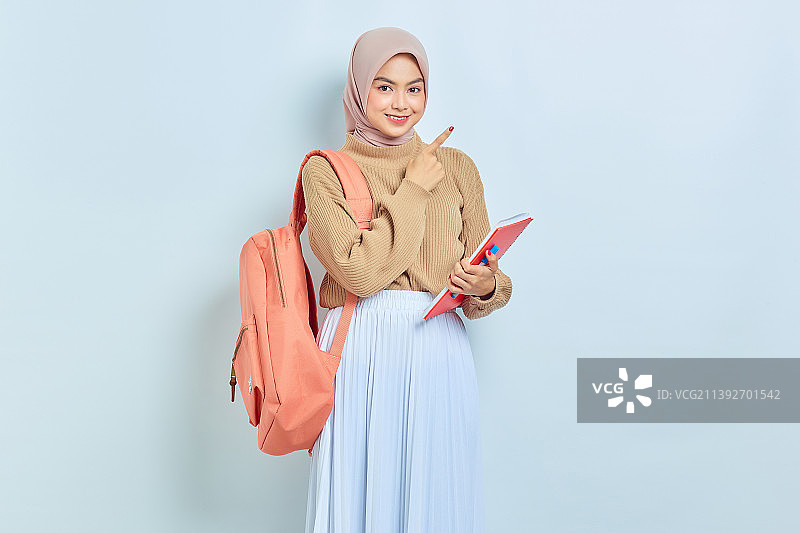 微笑的年轻亚洲穆斯林女学生，穿着棕色毛衣和背包，手指在一边图片素材