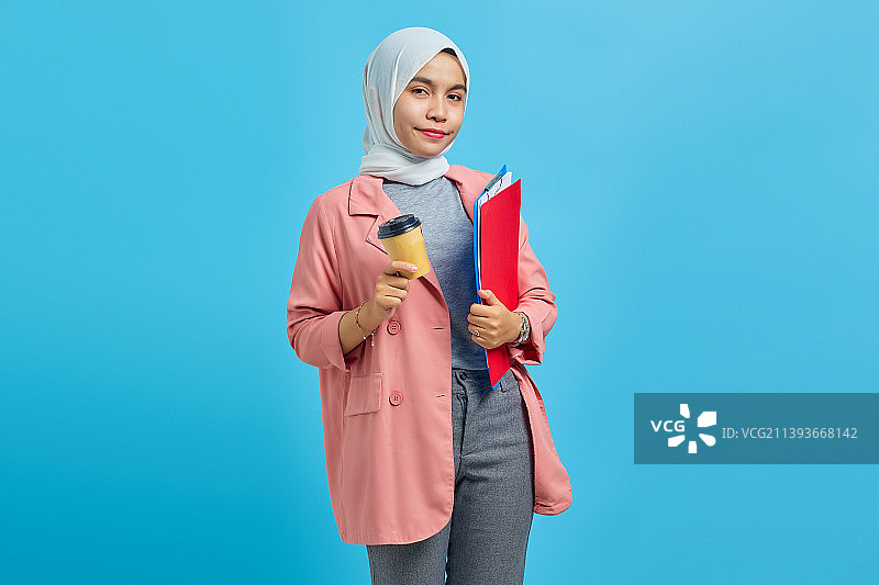 微笑的年轻亚洲妇女拿着文件夹和咖啡在蓝色的背景，印度尼西亚图片素材