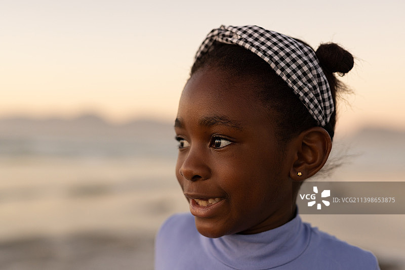 微笑的非洲裔美国可爱的女孩在日落时看着远处的大海和晴朗的天空的特写图片素材