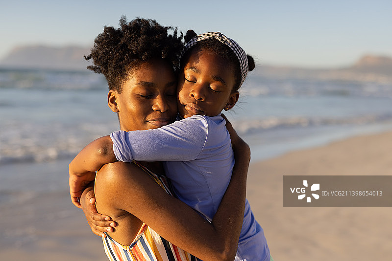 非洲裔美国年轻女子闭着眼睛，在日落时面对着大海和晴朗的天空拥抱女儿图片素材