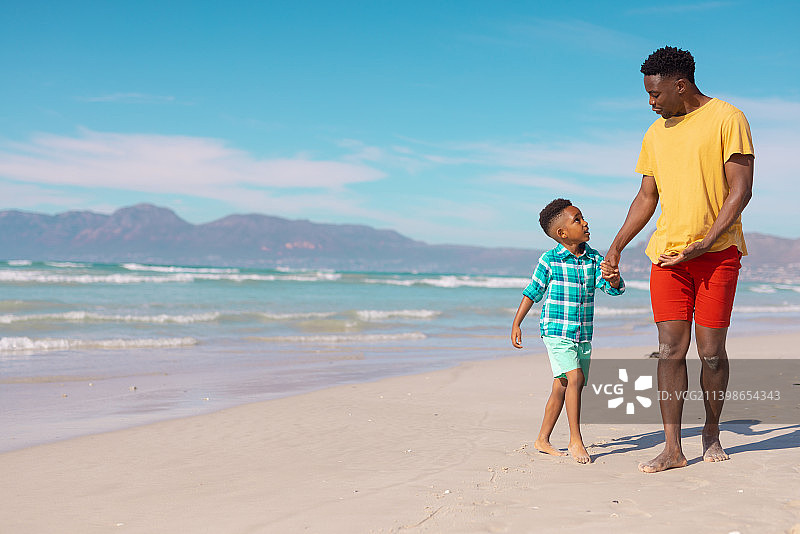 一个非洲裔美国男孩牵着年轻父亲的手，在蓝天下走在沙滩上图片素材