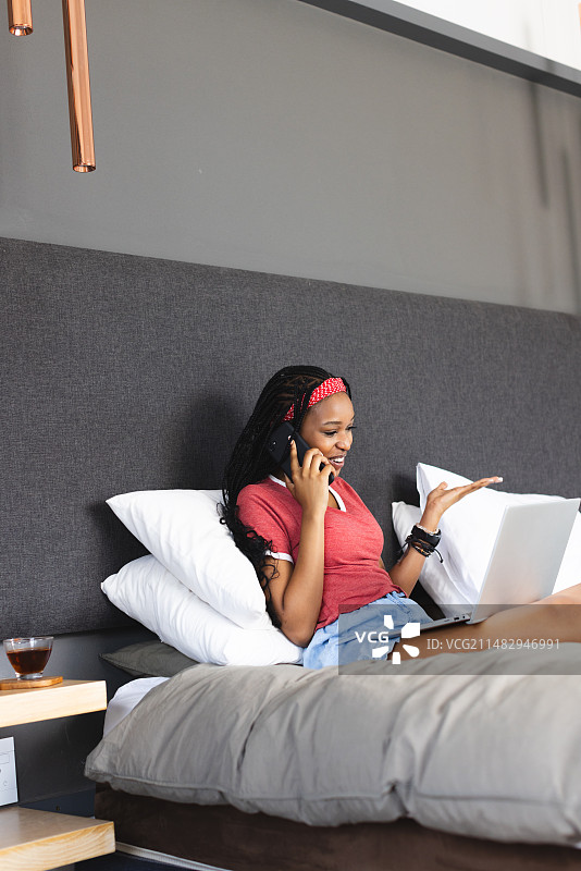 年轻的非洲裔美国妇女一边打电话，一边在床上用笔记本电脑复印图片素材
