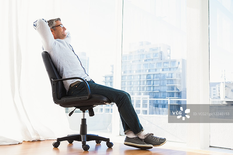 微笑着戴眼镜的男人坐在办公椅上，在公寓里放松图片素材