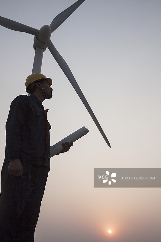 日落时分，一位年轻的男工程师手持蓝图，站在风力涡轮机旁图片素材