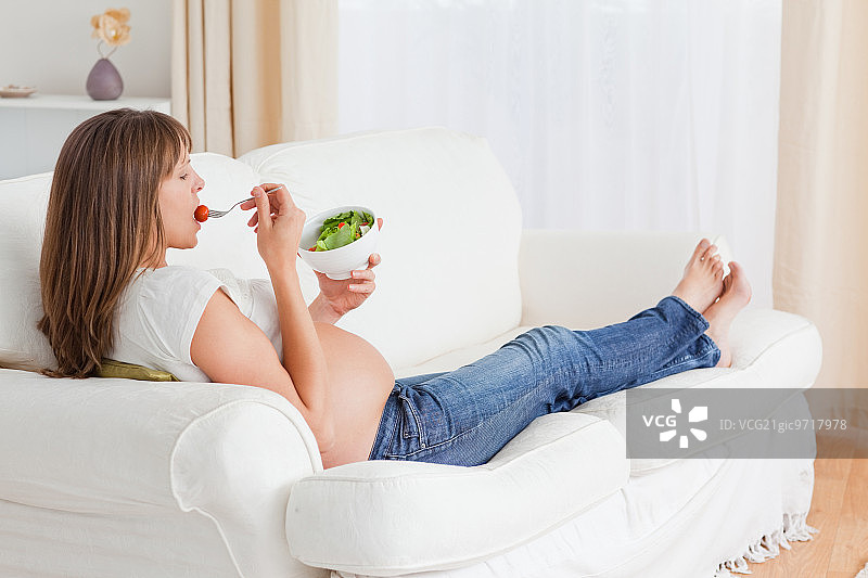 一个漂亮的孕妇躺在公寓的沙发上吃沙拉图片素材
