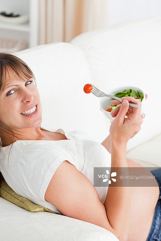 迷人的孕妇躺在公寓的沙发上吃沙拉图片素材