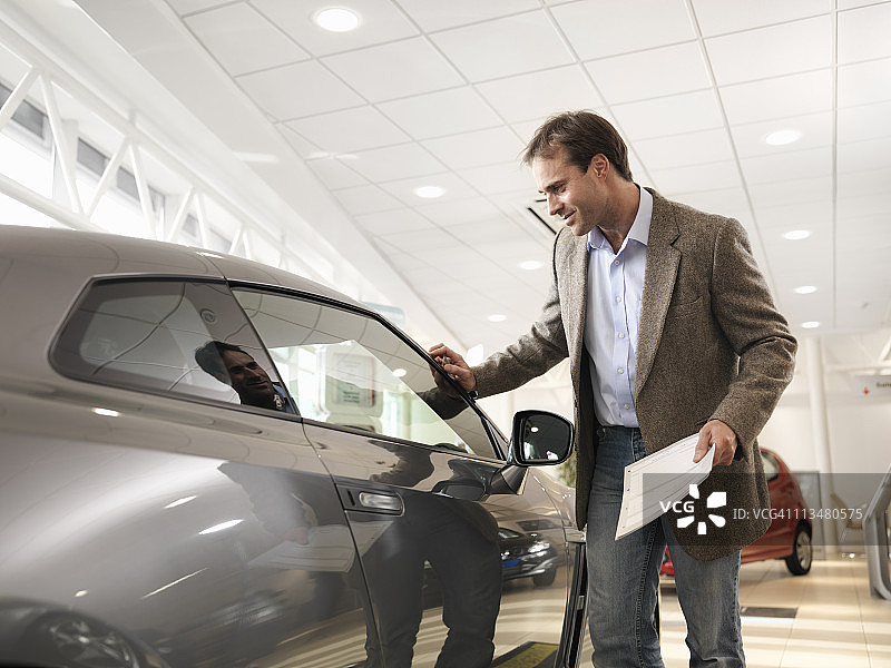 在汽车经销店里，男顾客拿着纸，用手摸着车看车图片素材