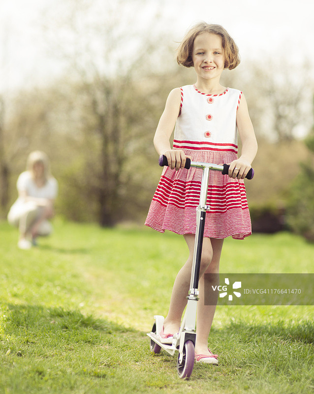 小女孩骑着滑板车穿过公园，后面是妈妈图片素材