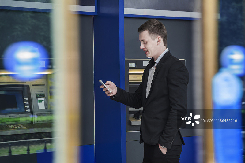 微笑的年轻商人站在自动取款机前，看着他的手机图片素材