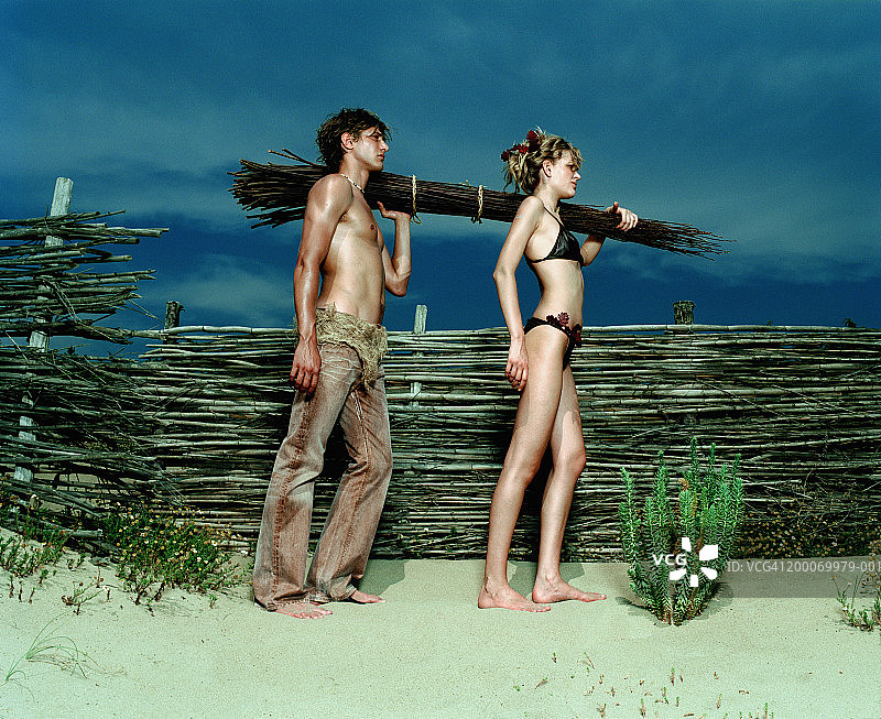 一名年轻男子和一名女子背着一捆木头在海滩上，侧视图片素材