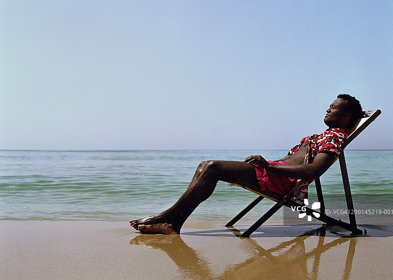 一个男人坐在海滩的躺椅上，闭着眼睛，侧视图片素材