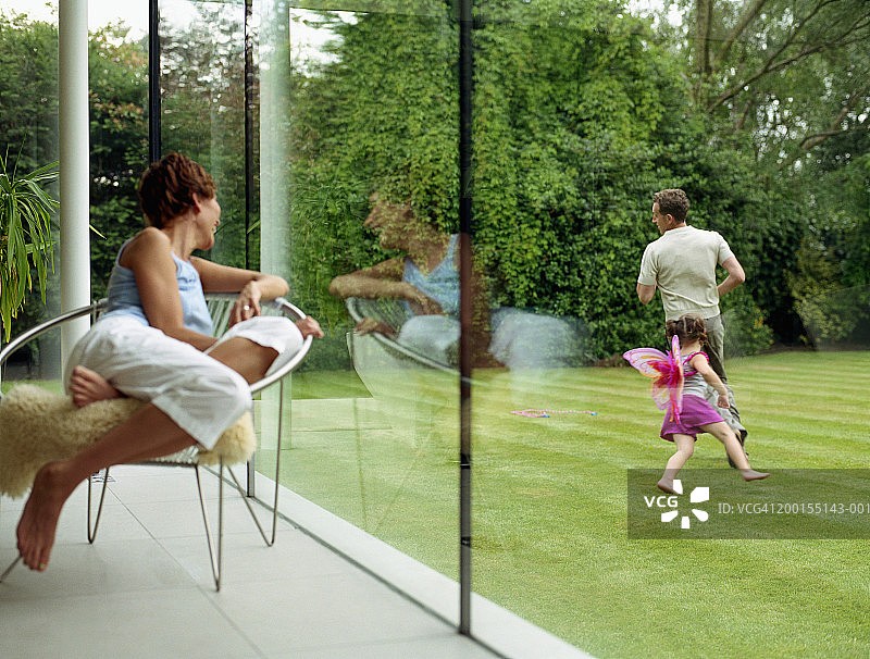 一个女人看着窗外的父亲和女儿在花园里奔跑图片素材